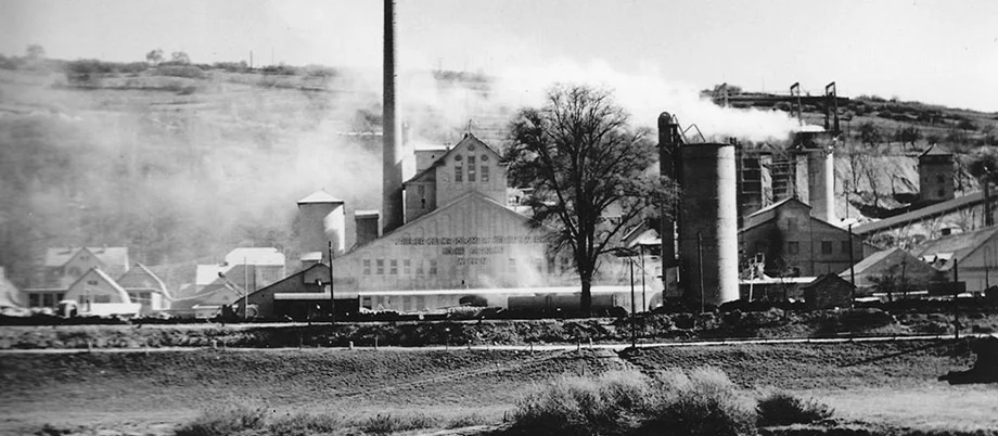 Image historique : les bâtiments de l’usine et leurs cheminées enveloppées d’une fumée blanche ; au premier plan : la Moselle